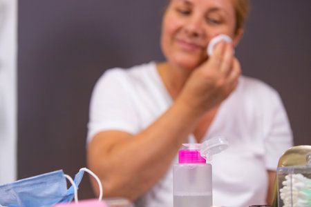 The pose of a woman with acne problem on the face having in front of her medical mask, bottle of lotion and cotton buds on the table, so she uses cotton disk to wipe her face.の写真素材
