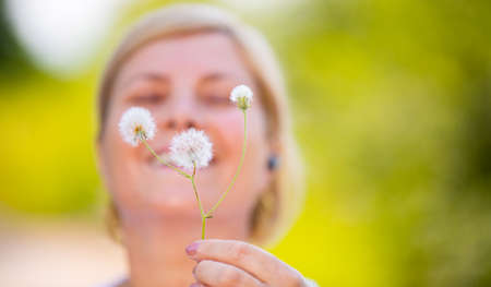 Close up photo of a happy woman blonde holding in front of the camera dandelion sprig being in nature to breath fresh air.の写真素材