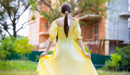 The back view photo of a gorgeous brunette young lady in beautiful yellow long dress holding it by the sides, looking as a princess.の写真素材
