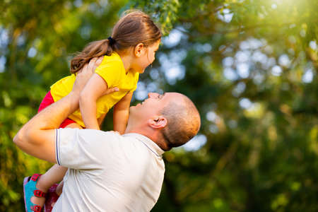 Father playing with his daughter in the park raising her up overhead bent his elbows and looking at her with love, daddy day came.の写真素材