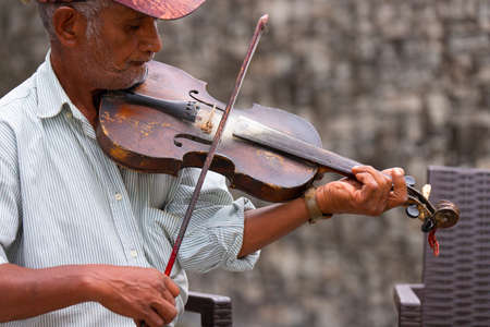 Profile photo of a senior adult man wearing a cap and striped t-shirt playing violin sitting on a chair outdoors, with his music he cheers up passers-by..の写真素材