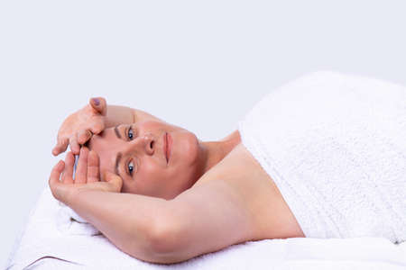 Calm relaxed woman lying on the massage table in a white towel keeping hands on her forehead waiting for a face massage procedures. ... High quality photoの写真素材