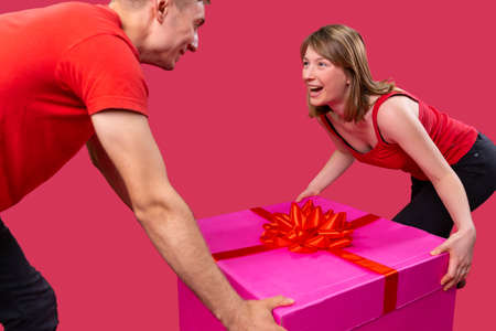 One nice lovely couple in red color t-shirts standing opposite each other and keeping on both sides the present with the red bow.の写真素材
