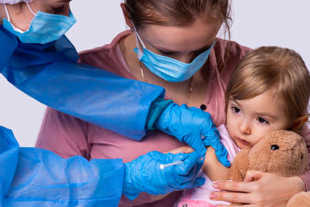 Close up photo of a mother in a face mask holding her little girl on her knees and the woman medic is putting a vaccine to her daughter using disposable syringe with needle. High quality photoの写真素材
