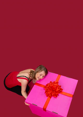 Smiling woman hugging the pink big present box with a huge red bow wearing t-shirt on shoulders straps.の写真素材