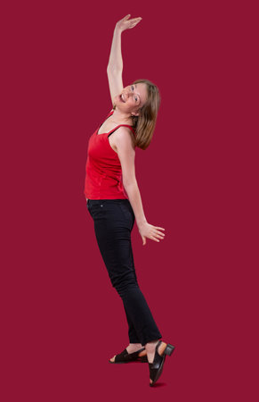 The photo of a joyful happy young woman in red t-shirt and black jeans posing on the camera with raised one hand up and another hand down.の写真素材