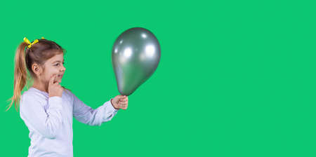 Adorable shiny young girl holding a dark green balloon in front of her and thinking to whom to give it.の写真素材