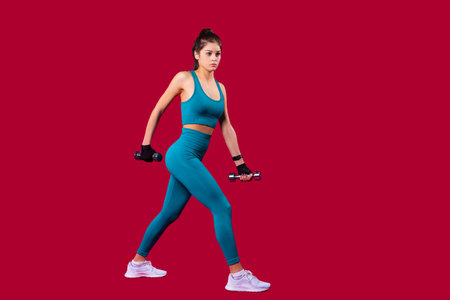 Young slim smiling woman in sports clothes taking a step forward with dumbbells holding un her hands doing fitness.の写真素材