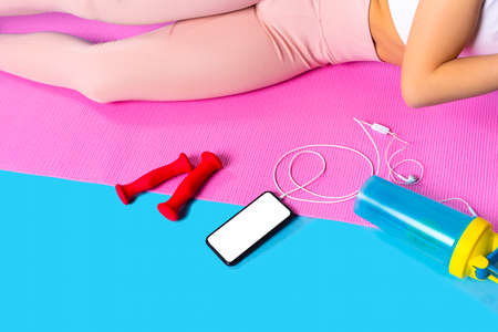 The photo of a lower body part of a fitness girl lying on the mat with red dumbbells, bottle of water and a phone near.の写真素材