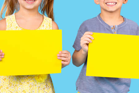 Happy boy in violet button t-shirt and girl in yellow floral dress holding two sheets of yellow color paper on the blue background.の写真素材