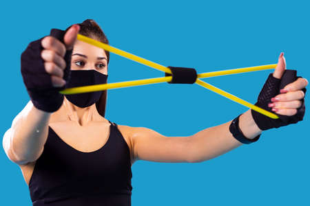 Close up of a fitness young girl using sports elastic rubber trying to stretch it wearing textile black mask in pandemic time.の写真素材
