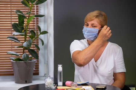 Caucasian woman blonde sitting at the table with cosmetics on it, tries to get off a medical mask to wipe her face with lotion after wearing the mask.の写真素材