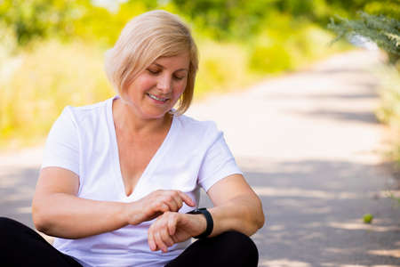 Close up photo of a sporty woman sitting with cross legs on the path in the forest pointing on her digital watch on her hand and is smiling.の写真素材