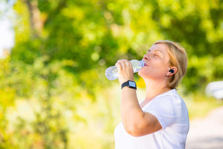 Beautiful sporty blonde woman wearing headphones and digital smart watch has a break after jogging outdoors in the park, drinking water from the plastic bottle.の写真素材