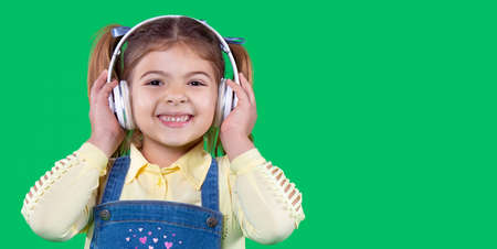 Cheerful small school aged girl in denim sundress holding her headphones on her head, listening the music and is very happy.の写真素材