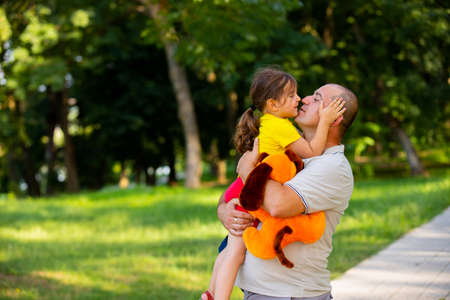 Loving and caring dad holding his daughter in his hands, hugging her keeping the dog soft toy in his hand and little girl holds her father head wants to give him a kiss.の写真素材