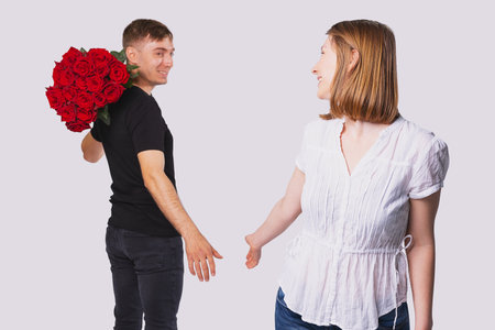 Young couple standing in the distance from each other, boyfriend holding a big bouquet of red roses on his shoulder to give to his blonde girlfriend.の写真素材