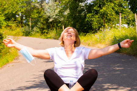 Joyful senior woman being in the park sitting on the road crossing legs holding hands on the sides enjoying a sunny day with the medical mask on one hand and digital watch on another hand.の写真素材