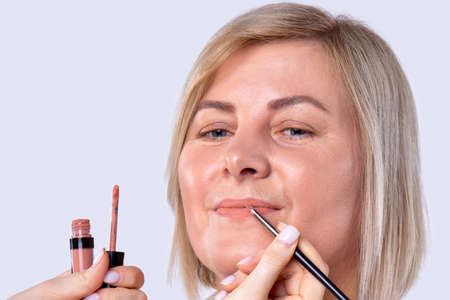 Visagist using lip gloss and lipstick brush applying makeup to a blonde pretty woman in a beauty salon for going out.の写真素材