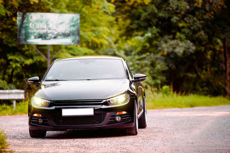 The black beautiful glossy car sedan rides on the road in the forest with no number and with headlights on.の写真素材