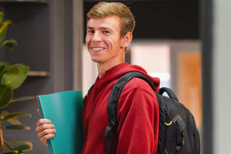 Side view photo of a happy student man in university wearing red hoodie with backpack on the back holding the color folder in his hand.の写真素材