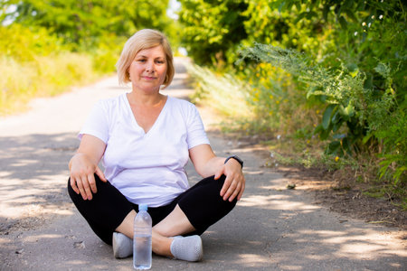 Pensive woman resting sitting on the road with cross legs in sports clothes keeping her hands on her knees, the bottle of water is near the woman.の写真素材