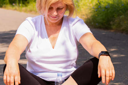 Happy confident senior blonde woman meditating in lotus position on the road in the park wearing smart watch on her hand and a bottle of water .の写真素材