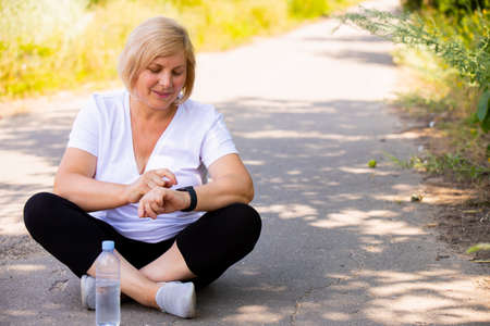 The photo of a happy blonde sporty woman sitting in a lotus position on the road in the park wearing sports clothes pointing on the digital watch on her hand.の写真素材