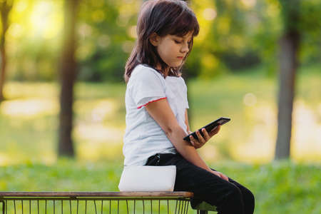 Side view photo of a pretty young girl sitting on a bench holding the phone in her hand and browsing interesting information.の写真素材