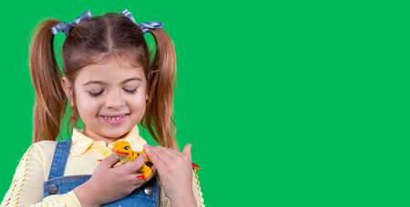 Banner, long format. Beautiful lovely girl with ponytails wearing denim sundress and yellow blouse is happy and holding in her arms a little toy lizard, empty space for the text.の写真素材