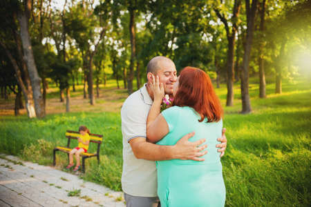 Beautiful loving couple of pregnant woman and her husband standing together on the path in the park embracing and kissing, their little daughter is sitting on a bench.の写真素材