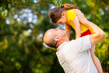 Loving father took his little daughter in his arms and raised her up, they are looking very happy together spending splendid sunny day in the park.の写真素材