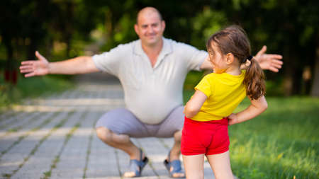 Happy dad squatting down on his knees is smiling and he wants to embrace his daughter with love, spend good time together in the park.の写真素材