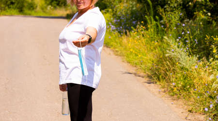Cropped photo of a fitness woman standing on the road in the forest holding the bottle of water and medical mask in her hands.の写真素材