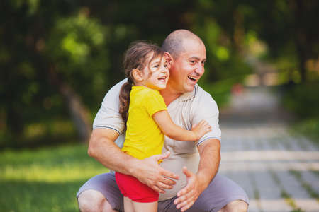 Close up photo of a nice daughter in yellow t-shirt and red shorts laughing with her daddy and he is embracing her outdoors.の写真素材