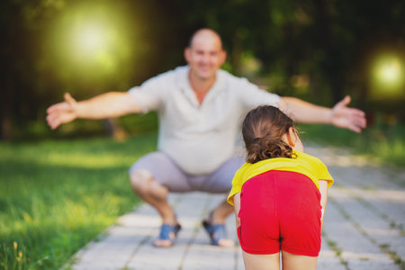 Back view photo of a little girl tilted her torso forward ready to run into the arms of her daddy squatting outdoors.の写真素材