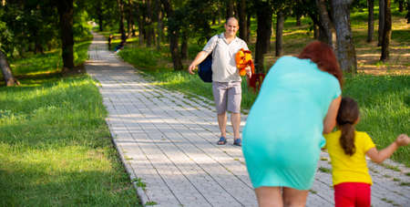 Mother and little daughter meeting daddy on the path in the forest, he is holding a big bag and a soft dog toy for his little girl.の写真素材
