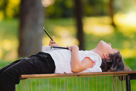 Close up photo of a young girl lying on a bench on her back in the park holding the smart phone and small purse in her hands, resting after voluntary work.の写真素材