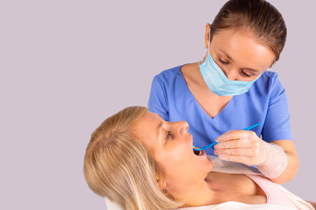 Blonde woman sitting in a chair in the dentist office and woman stomatologist in a medical mask and protective gloves examines the oral cavity.の写真素材