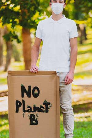 Full-length photo of a young man in a medical mask standing in the park with cardboard poster No Planet B inscription, protests to save planet earth.の写真素材