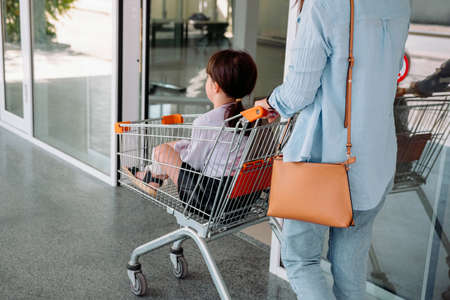 Back view photo of a school aged girl sitting in the grocery cart and her mother pushes the cart to enter the store.の写真素材