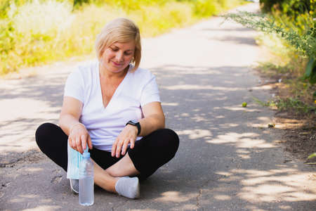 Fitness happy woman sitting in the lotus position on the road in the park single with a bottle of water down, keeping medical mask on her hand.の写真素材