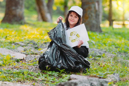 Close of photo of a pretty young girl sitting on the ground in the park holding a paper with a recycle sign and gather it into the garbage bag.の写真素材