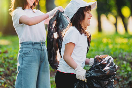 Side view photo of a happy young girl in white cap and t-shirt wearing gloves keeping the garbage bag for the rubbish and other girl standing near holding the garbage bag, to keep the nature clean.の写真素材