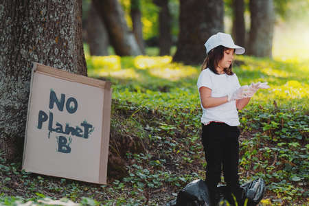 Side view photo of a young volunteer girl taking on protective gloves ready for gathering rubbish in the park behind her there is a cardboard near the tree with inscription No Planet B.の写真素材