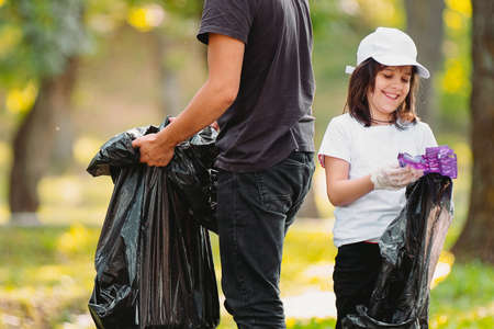 Picture of a smiling beautiful girl in protective gloves and adult man volunteer gathering plastic rubbish together outdoors using big black garbage bags.の写真素材