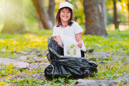 Close up photo of a nice young girl outdoors having wide smiling on her face, sitting on the ground garbage bag and a paper with a recycle sign on it.の写真素材