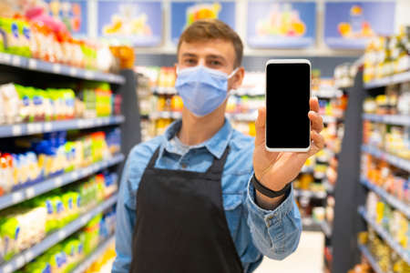 Young store worker man with surgical medical mask in black apron showing the smart phone display on store market background.の写真素材