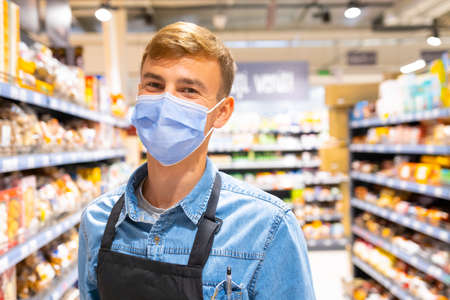 Close up photo of a happy man store worker in a medical mask wearing denim shirt having pen in his chest pocket and black apron standing among the shelves with the goods.の写真素材