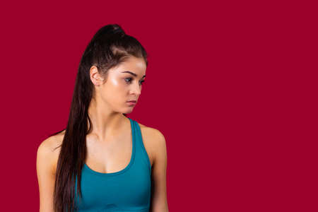 The profile photo of a sporty young girl in blue top and long brunette ponytail tired after training, poses over the red background in the studio.の写真素材